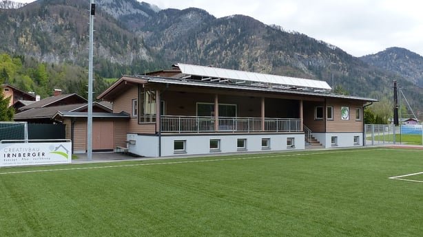 Außenansicht des teilsolar beheizten Vereinsheims des USV Scheffau. Das Gebäude mit Holzfassade und Solaranlage auf dem Dach liegt direkt am Fußballplatz vor einer Bergkulisse.