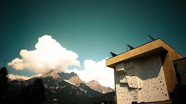 Künstlerische Aufnahme des Kletterturms der solar beheizten Kletterhalle Saalfelden vor einer imposanten Bergkulisse.