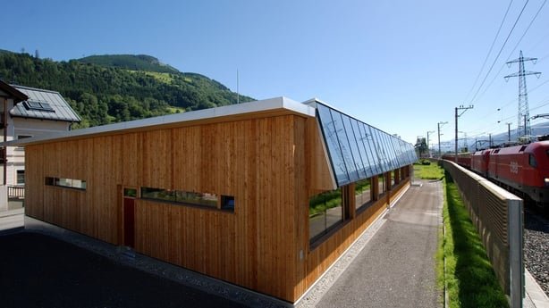 Außenansicht der vollsolar beheizten Tischlerei-Lehrwerkstätte in Bruck. Das moderne Holzgebäude mit integrierten Solarkollektoren in der Fassade steht direkt neben den Bahngleisen.